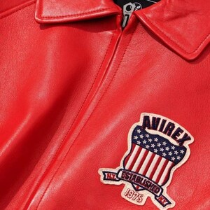 May include: Close-up of a red leather jacket with a collar and zipper. A patch on the jacket features the word "AVIREX" above a design of the American flag and the text "NY ESTABLISHED 1975 NY".