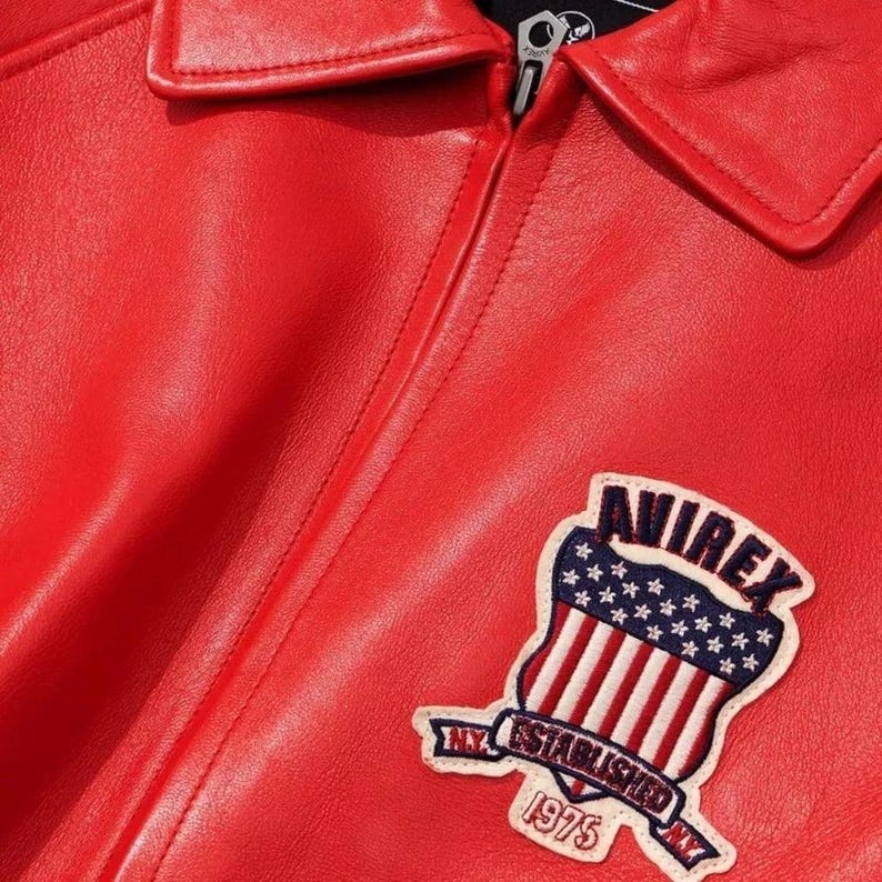 May include: Close-up of a red leather jacket with a collar and zipper. A patch on the jacket features the word "AVIREX" above a design of the American flag and the text "NY ESTABLISHED 1975 NY".