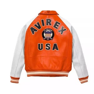Könnte beinhalten: Orange-weiße Lederjacke mit dem Wort "AVIREX" oben und "USA" unten. Die Jacke hat weiße Ärmel und einen gerippten Kragen und Bündchen. Ein Aufnäher mit den Worten "ALL AMERICANS ESTABLISHED 1975" befindet sich in der Mitte.