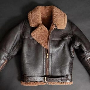 Puede incluir: Chaqueta de cuero marrón oscuro con cuello y forro de borrego. La chaqueta tiene cremallera frontal, mangas con cremallera y cinturón en la cintura. El exterior de la chaqueta es de color marrón oscuro y el borrego es de color marrón claro.