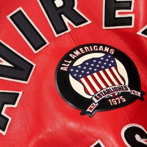 May include: Close-up of a red leather jacket with white and black lettering spelling out "AIR" and "RE". A circular patch reads "ALL AMERICANS ESTABLISHED 1975 N.Y." with an American flag design.