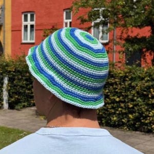 May include: A striped bucket hat in shades of blue, green, and white. The hat is crocheted and has a rounded crown and a short brim. The text "THE STRIPED BUCKET HAT" is at the top of the image, with "By Josephinejar" below it.