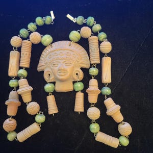 May include: A necklace featuring a central terracotta-colored pendant shaped like a human face with a headdress. The necklace is strung with alternating light green marbled beads and textured tan cylindrical and spherical clay-like beads.
