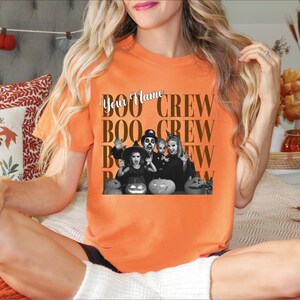 K&ouml;nnte beinhalten: Ein orangefarbenes Kurzarm-T-Shirt mit einer schwarz-wei&szlig;en Grafik einer Familie in Halloween-Kost&uuml;men mit K&uuml;rbissen. Der Text "Your Name" ist in Schreibschrift &uuml;ber dem wiederholten braunen Text "BOO CREW" in Blockschrift gedruckt.