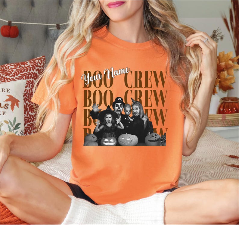 K&ouml;nnte beinhalten: Ein orangefarbenes Kurzarm-T-Shirt mit einer schwarz-wei&szlig;en Grafik einer Familie in Halloween-Kost&uuml;men mit K&uuml;rbissen. Der Text "Your Name" ist in Schreibschrift &uuml;ber dem wiederholten braunen Text "BOO CREW" in Blockschrift gedruckt.