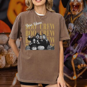 K&ouml;nnte beinhalten: Ein braunes Kurzarm-T-Shirt mit einem Grafikdruck, der den Text "Your Name" und "BOO CREW" in goldener Schrift wiederholt. Die Grafik zeigt ein schwarz-wei&szlig;es Bild einer Familie in Kost&uuml;men mit K&uuml;rbissen im Vordergrund.