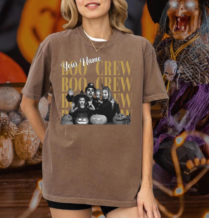 K&ouml;nnte beinhalten: Ein braunes Kurzarm-T-Shirt mit einem Grafikdruck, der den Text "Your Name" und "BOO CREW" in goldener Schrift wiederholt. Die Grafik zeigt ein schwarz-wei&szlig;es Bild einer Familie in Kost&uuml;men mit K&uuml;rbissen im Vordergrund.