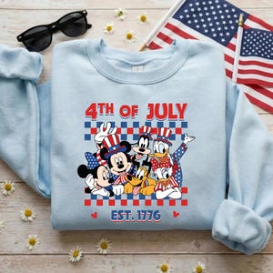 Pu&ograve; includere: Una felpa girocollo azzurro chiaro con una grafica di Topolino, Minni, Pippo, Paperino e Pluto che indossano cappelli patriottici. Il testo 4TH OF JULY e EST. 1776 &egrave; posizionato sopra e sotto un motivo a scacchi rosso, bianco e blu.