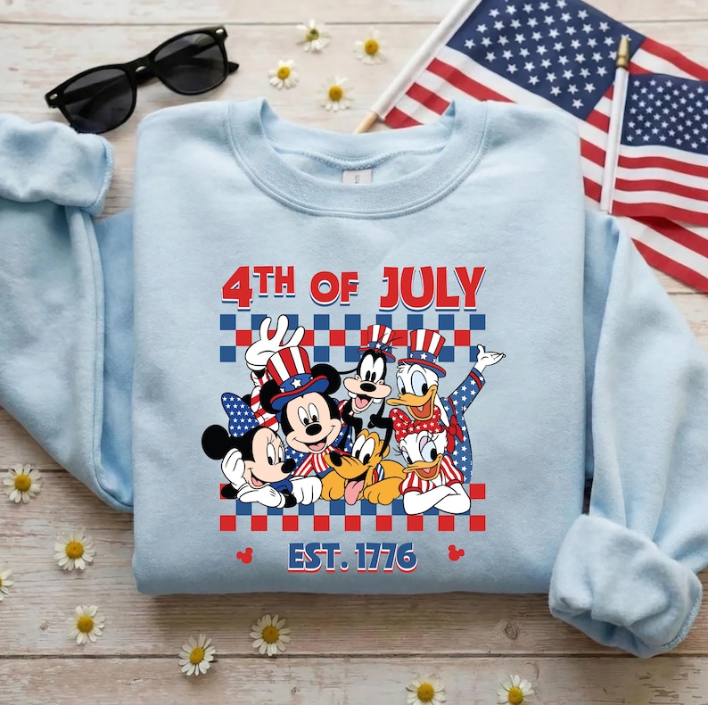 Pu&ograve; includere: Una felpa girocollo azzurro chiaro con una grafica di Topolino, Minni, Pippo, Paperino e Pluto che indossano cappelli patriottici. Il testo 4TH OF JULY e EST. 1776 &egrave; posizionato sopra e sotto un motivo a scacchi rosso, bianco e blu.