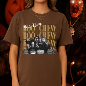 K&ouml;nnte beinhalten: Ein braunes T-Shirt mit kurzen &Auml;rmeln und einem Grafikdruck, der den Text "Your Name" &uuml;ber den in einem Raster wiederholten W&ouml;rtern "BOO CREW" zeigt. Das Design enth&auml;lt ein Schwarz-Wei&szlig;-Foto einer Familie in Halloween-Kost&uuml;men.