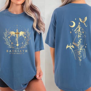 a woman wearing a blue t-shirt with a design featuring a dragon, a crescent moon, and a cross. The design is gold-colored and appears to be a symbol or emblem associated with the "Bagith" brand.