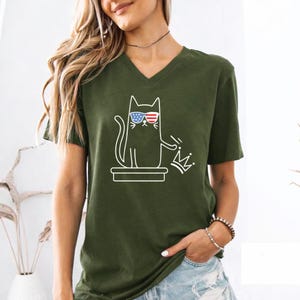 May include: Olive green V-neck t-shirt featuring a white graphic of a cat wearing sunglasses with an American flag design. The cat is holding a crown. The shirt is a casual style.