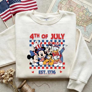 Pu&ograve; includere: Una felpa girocollo color crema con una grafica di Topolino, Minni, Pippo, Paperino, Paperina e Pluto che indossano cappelli patriottici. Il testo recita 4TH OF JULY e EST. 1776 su uno sfondo a scacchi rossi e blu.