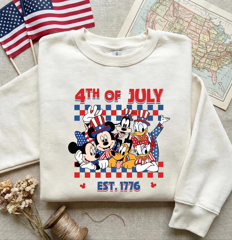 Pu&ograve; includere: Una felpa girocollo color crema con una grafica di Topolino, Minni, Pippo, Paperino, Paperina e Pluto che indossano cappelli patriottici. Il testo recita 4TH OF JULY e EST. 1776 su uno sfondo a scacchi rossi e blu.