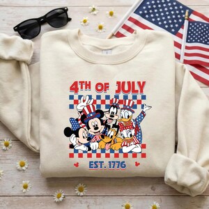 Pu&ograve; includere: Una felpa girocollo color crema con una grafica di Topolino, Minni, Pippo, Pluto, Paperino e Paperina che indossano cappelli patriottici. Il testo recita 4TH OF JULY e EST. 1776 su uno sfondo a scacchi rosso, bianco e blu.