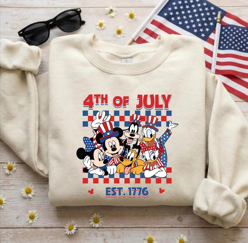 Pu&ograve; includere: Una felpa girocollo color crema con una grafica di Topolino, Minni, Pippo, Pluto, Paperino e Paperina che indossano cappelli patriottici. Il testo recita 4TH OF JULY e EST. 1776 su uno sfondo a scacchi rosso, bianco e blu.