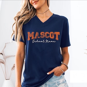 May include: A navy blue V-neck t-shirt featuring the word MASCOT in distressed orange block lettering with the text School Name written in a white cursive script font centered directly underneath the primary graphic on the chest area.