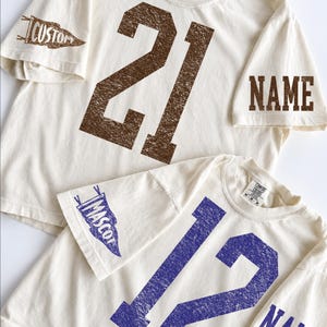 May include: Two off-white short-sleeve t-shirts with distressed-style graphics. The top shirt features the number 21 in brown, a pennant graphic with the word CUSTOM, and the word NAME. The bottom shirt features the number 12 in purple, a pennant graphic with the word MASCOT, and the word NAME.