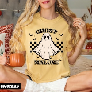 May include: A mustard yellow short-sleeve t-shirt featuring a graphic of a ghost with facial tattoos, positioned above a black and white checkered pattern. The text GHOST MALONE is printed in a bold, dark font, surrounded by small bats and stars.
