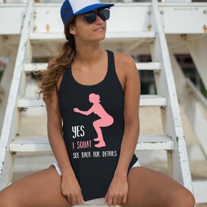 May include: A black racerback tank top featuring a pink silhouette graphic of a person performing a squat. The text printed in white and pink reads: YES I SQUAT SEE BACK FOR DETAILS. The garment has a scoop neckline and a sleeveless design.