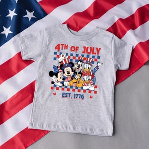 以下が含まれることがあります： ヘザーグレーの半袖Tシャツ。ミッキーマウス、ミニーマウス、グーフィー、プルート、ドナルドダック、デイジーダックが愛国的な帽子をかぶったグラフィックが描かれています。キャラクターの上下には「4TH OF JULY」と「EST. 1776」の文字があります。