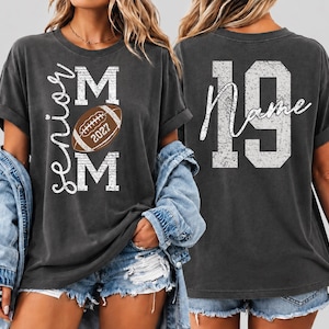 May include: A charcoal gray t-shirt featuring a front graphic with the text Senior MOM and a brown football with the year 2027. The back of the shirt displays the large number 19 in a distressed white font with the word Name written in script.