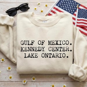 May include: A cream-colored crewneck sweatshirt featuring black text that reads GULF OF MEXICO. KENNEDY CENTER. LAKE ONTARIO. The garment has long sleeves with ribbed cuffs and a ribbed neckline, presented alongside black sunglasses and a small flag.