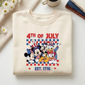 Pu&ograve; includere: Una felpa girocollo color crema con una stampa grafica di Topolino, Minni, Pippo, Paperino, Paperina e Pluto che indossano abiti patriottici. Il testo recita 4TH OF JULY e EST. 1776 sopra un motivo a scacchi rosso e blu.