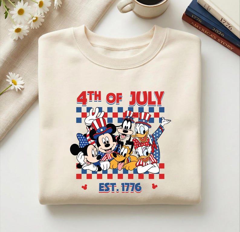 Pu&ograve; includere: Una felpa girocollo color crema con una stampa grafica di Topolino, Minni, Pippo, Paperino, Paperina e Pluto che indossano abiti patriottici. Il testo recita 4TH OF JULY e EST. 1776 sopra un motivo a scacchi rosso e blu.
