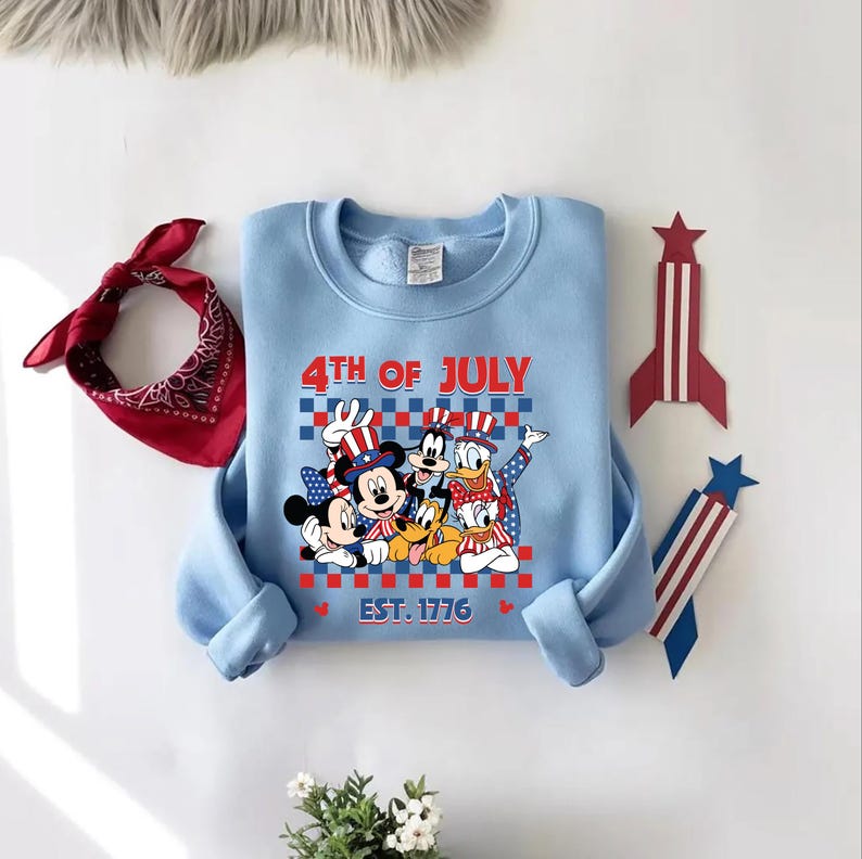 Pu&ograve; includere: Una felpa girocollo azzurro chiaro con una grafica di personaggi dei cartoni animati che indossano cappelli patriottici. Il testo recita 4TH OF JULY e EST. 1776. Una bandana rossa a motivi e due decorazioni a razzo sono poste attorno.