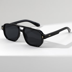 May include: Black aviator-style sunglasses with dark lenses. The sunglasses have a double bridge and thick black frames. Small gold-colored accents are visible on the frame. The temples are also black.