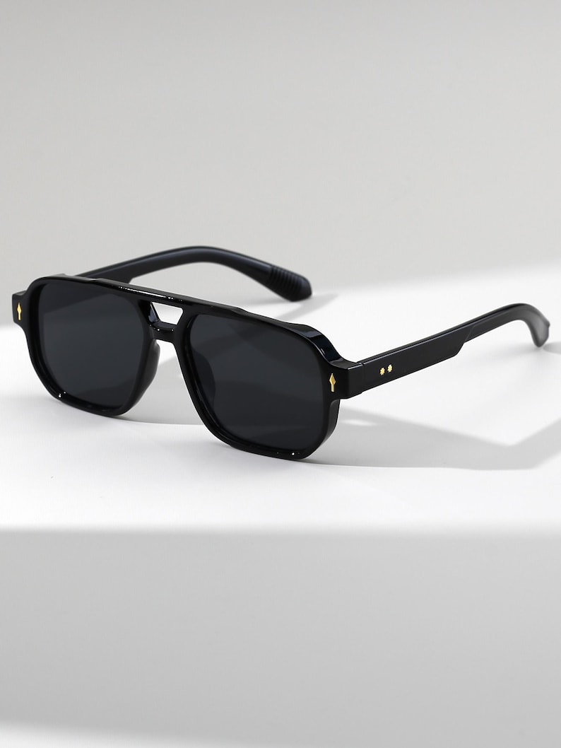 May include: Black aviator-style sunglasses with dark lenses. The sunglasses have a double bridge and thick black frames. Small gold-colored accents are visible on the frame. The temples are also black.
