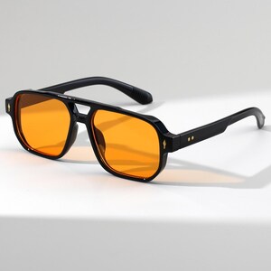 May include: Black sunglasses with orange lenses. The sunglasses have a square frame with a double bridge and a thick black frame. The lenses are a bright orange color. The arms of the sunglasses are black and have a slight curve.
