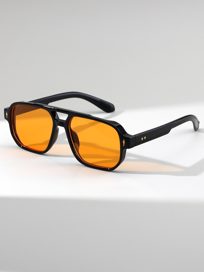 May include: Black sunglasses with orange lenses. The sunglasses have a square frame with a double bridge and a thick black frame. The lenses are a bright orange color. The arms of the sunglasses are black and have a slight curve.