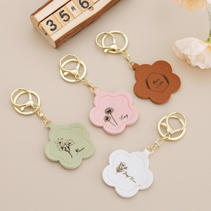Puede incluir: Un conjunto de llaveros con forma de flor en varios colores: marrón, rosa, verde y blanco. Cada llavero tiene un anillo y un cierre dorados. Los llaveros están personalizados con nombres y diseños florales.