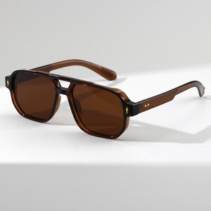 May include: Brown aviator sunglasses with a double bridge and dark lenses. The sunglasses have a square shape and a subtle gold accent on the temples. The frames and arms are a matching brown color.