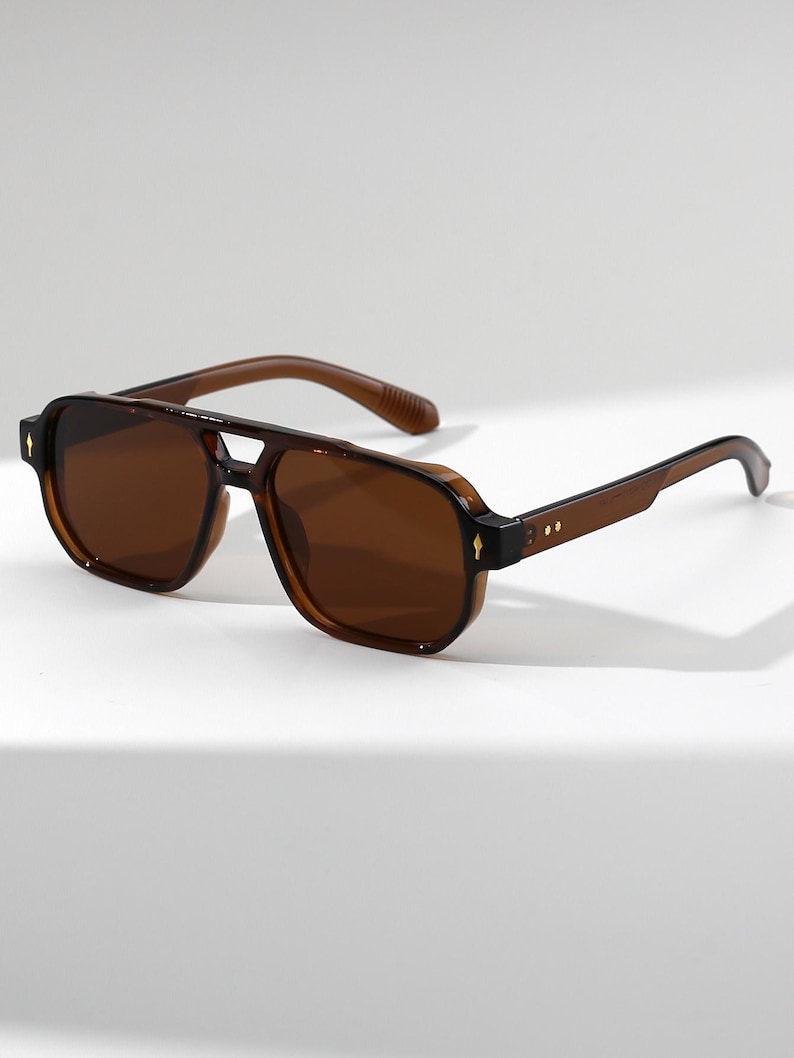 May include: Brown aviator sunglasses with a double bridge and dark lenses. The sunglasses have a square shape and a subtle gold accent on the temples. The frames and arms are a matching brown color.