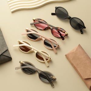 Op de afbeelding: Een assortiment zonnebrillen in verschillende kleuren en stijlen, waaronder zwart, schildpad en crème, met bijpassende etuis. De zonnebrillen hebben ovale monturen en getinte glazen, gerangschikt op een neutrale ondergrond.