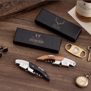 May include: A collection of engraved gifts, featuring a black box with a stag design and the name "Lucas", a box with "Groomsman" text, cufflinks, and a pocket watch. Also, two corkscrews, a cigar cutter, and decorative keys.