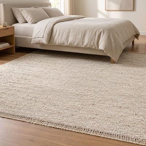 May include: A rectangular area rug featuring a thick, woven texture in a neutral beige and cream color palette. The rug is constructed with a chunky, looped yarn design and finished with short, straight fringe detailing along the shorter edges.