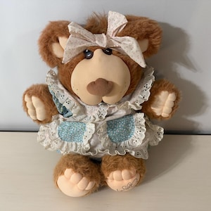 Op de afbeelding: Een bruine teddybeer met een witte en blauwe jurk en een bijpassende strik. De beer heeft een lichtbruine snuit en poten. De jurk heeft een blauw bloemenpatroon en witte kanten rand. De beer zit op een wit oppervlak.