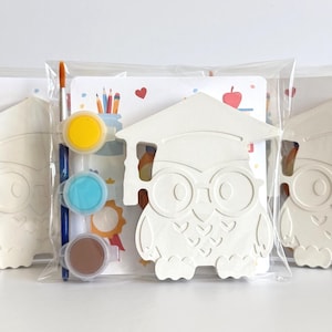 Op de afbeelding: Drie doe-het-zelf-schilder-uilen-sets. Elke set bevat een witte uilvormige figuur met een afstudeerhoed, verfpotjes in geel, blauw en bruin en een penseel. De sets zijn verpakt in doorzichtige plastic zakken.