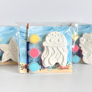 May include: Three individual craft kits in clear plastic packaging, each featuring a white plaster sea creature figure, a small paintbrush, and three small paint pots in yellow, pink, and blue, set against a printed underwater background.