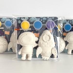 Puede incluir: Kits de manualidades de temática espacial para pintar. Cada kit incluye una figura blanca de astronauta, cohete o planeta, botes de pintura amarilla, azul y morada, y un pincel. Los kits están empaquetados en bolsas de plástico transparente.