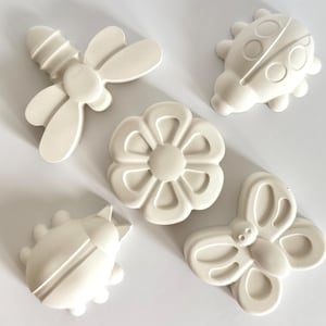 Puede incluir: Cinco formas decorativas de color blanquecino que representan insectos y una flor. Las formas incluyen una libélula, una mariquita, una mariposa, una flor y un escarabajo. Los objetos son de material sólido, sobre una superficie blanca.