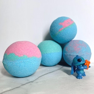 Könnte beinhalten: Mehrere bunte Badebomben in Blau-, Rosa- und Türkistönen sind auf einer weißen Oberfläche angeordnet. Eine kleine blaue Figur einer Zeichentrickfigur hält ein kleines orangefarbenes Geschenk. Die Badebomben haben ein strukturiertes, körniges Aussehen.