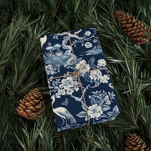 Pode incluir: Um presente embrulhado com um padrão floral e de pássaros azul marinho, amarrado com barbante. O papel de embrulho apresenta flores brancas, pássaros e elementos de paisagem. O presente é cercado por agulhas de pinheiro e pinhas.