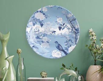 May include: A round decorative plate with a blue and white floral and bird design. The plate features a scenic landscape with a bird perched on a branch. The plate is displayed on a wooden surface with other decorative items.