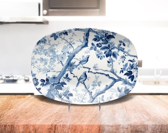 May include: Oval serving platter with a blue and white floral and bird design. The ceramic platter features a detailed illustration of branches, flowers, and two birds. The platter is displayed on a clear stand, showcasing its decorative pattern.