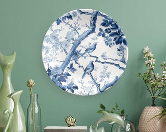 May include: A round, white decorative plate with a blue floral and bird design. The plate features a detailed illustration of birds perched on branches with leaves and flowers. The plate is mounted on a green wall.